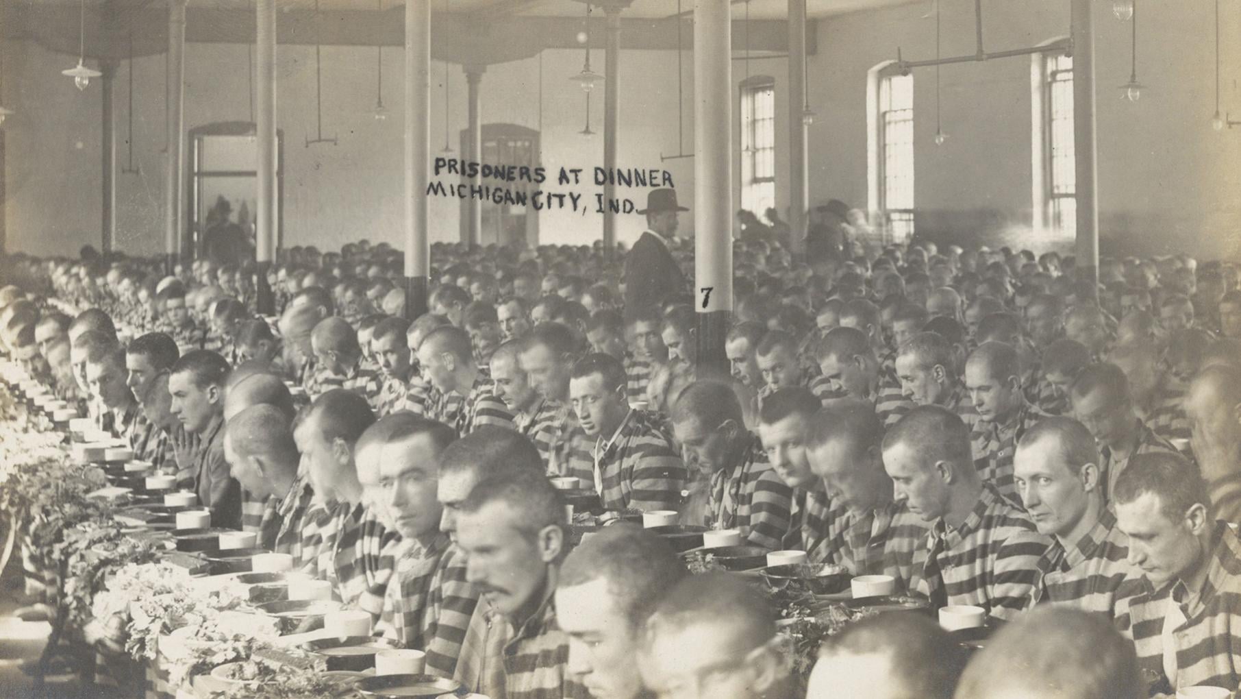Rows of prisoners in black and white stripes sit at long tables in a black and white photo