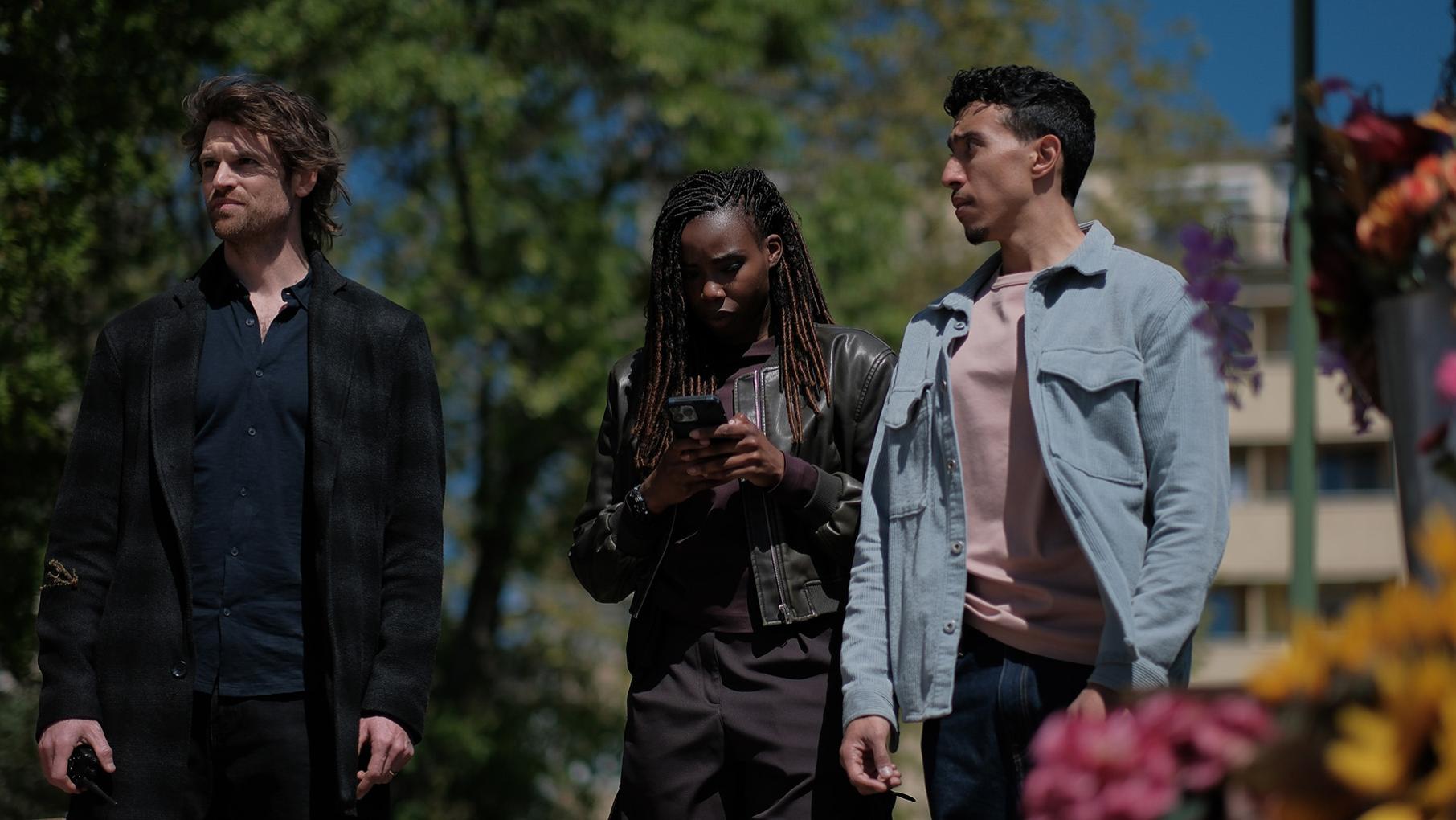 Janvier looks at her phone while Maigret and Lapointe stand on either side of her outside