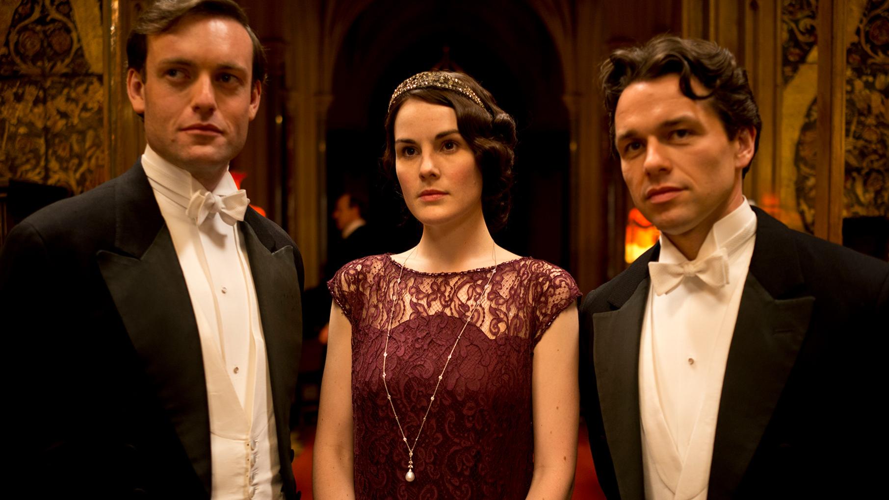 Mary stands in a maroon dress between Evelyn Napier and Charles Blake, who are in tuxedoes 