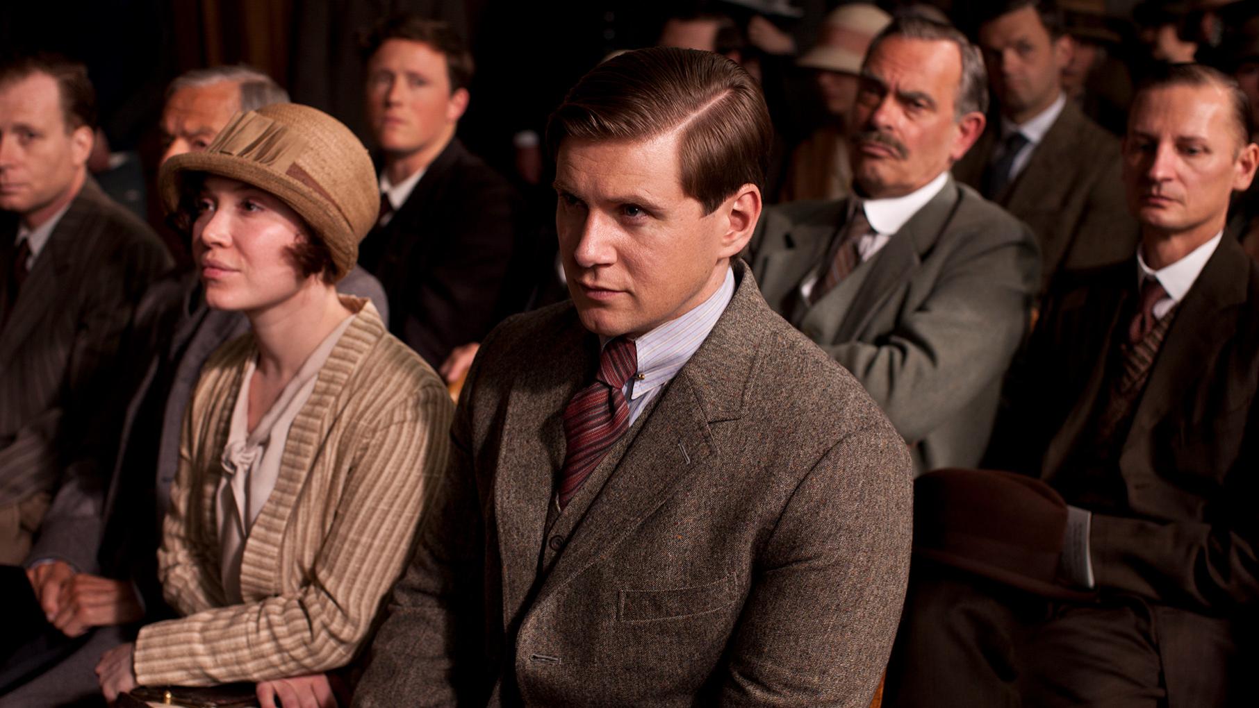 Tom sits in a suite and tie amongst other people in dark clothing