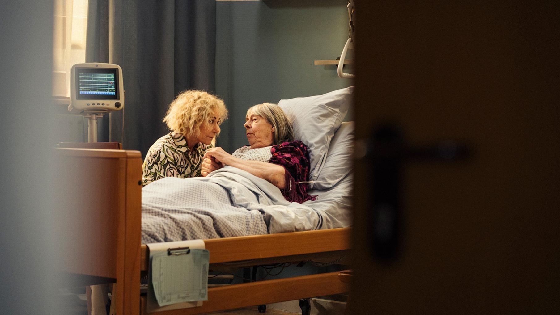Zelda leans over Adelaide in a hospital bed, seen through a partially closed dooer