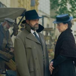 Oskar, with his collar turned up and a hat on, speaks to Therese in the street
