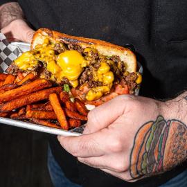A sandwich with a cheese sauce and sweet potato fries arranged on a try carried by a person with a hamburger tattoo on the back of his hand.