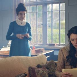 Cassy reads from pages on a couch while Jane stands expectantly behind
