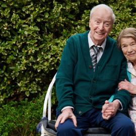Michael Caine and Glenda Jackson sit on a bench outside smiling