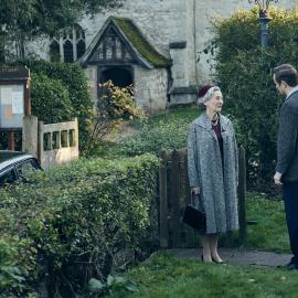 Daniel stands talking to his mother next to a hedge in front of the church