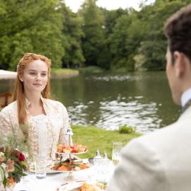 Irene sits at a table outside by a river facing Soames