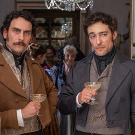 Fernand and Danglars stand in a doorway with glasses of champagne