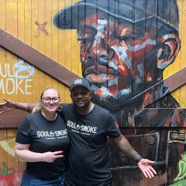 Heather Bublick and D'Andre Carter pose in front of a barn door painted with produce and a man in a cap
