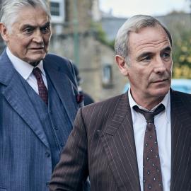 Jack and Geordie stand looking around the corner of a building in suits