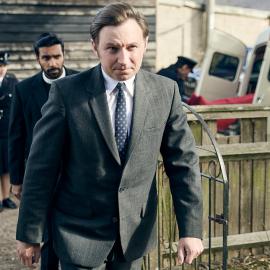 Larry marches through a yard with Alphy and policemen behind him