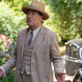 Atticus Pünd looks surprised standing outside in a hat and suit in front of a car