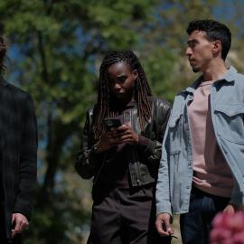 Janvier looks at her phone while Maigret and Lapointe stand on either side of her outside