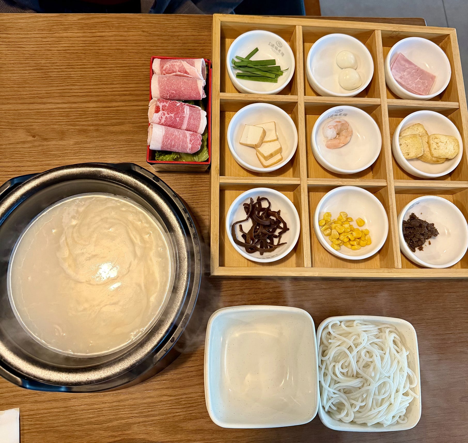 A bowl of soup next to dishes of meat, vegetables, and other garnishes and noodles